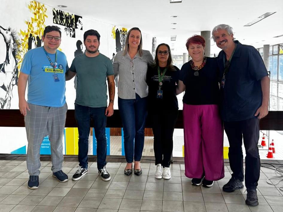 inserir aqui Grupo de seis profissionais reunidos em corredor com mural artístico moderno em preto e amarelo ao fundo, ambiente institucional iluminado.
