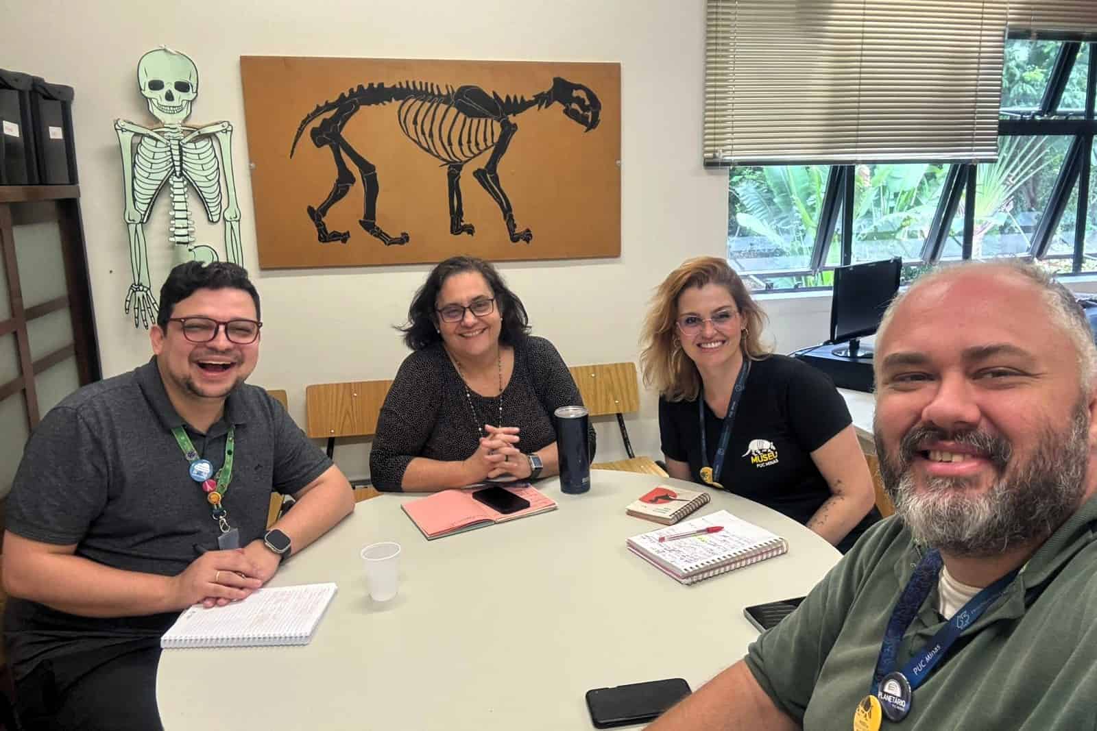 inserir aqui Quatro pessoas sorrindo para a câmera em evento CSM Minas. Ao fundo, auditório da UFMG com público e palco.