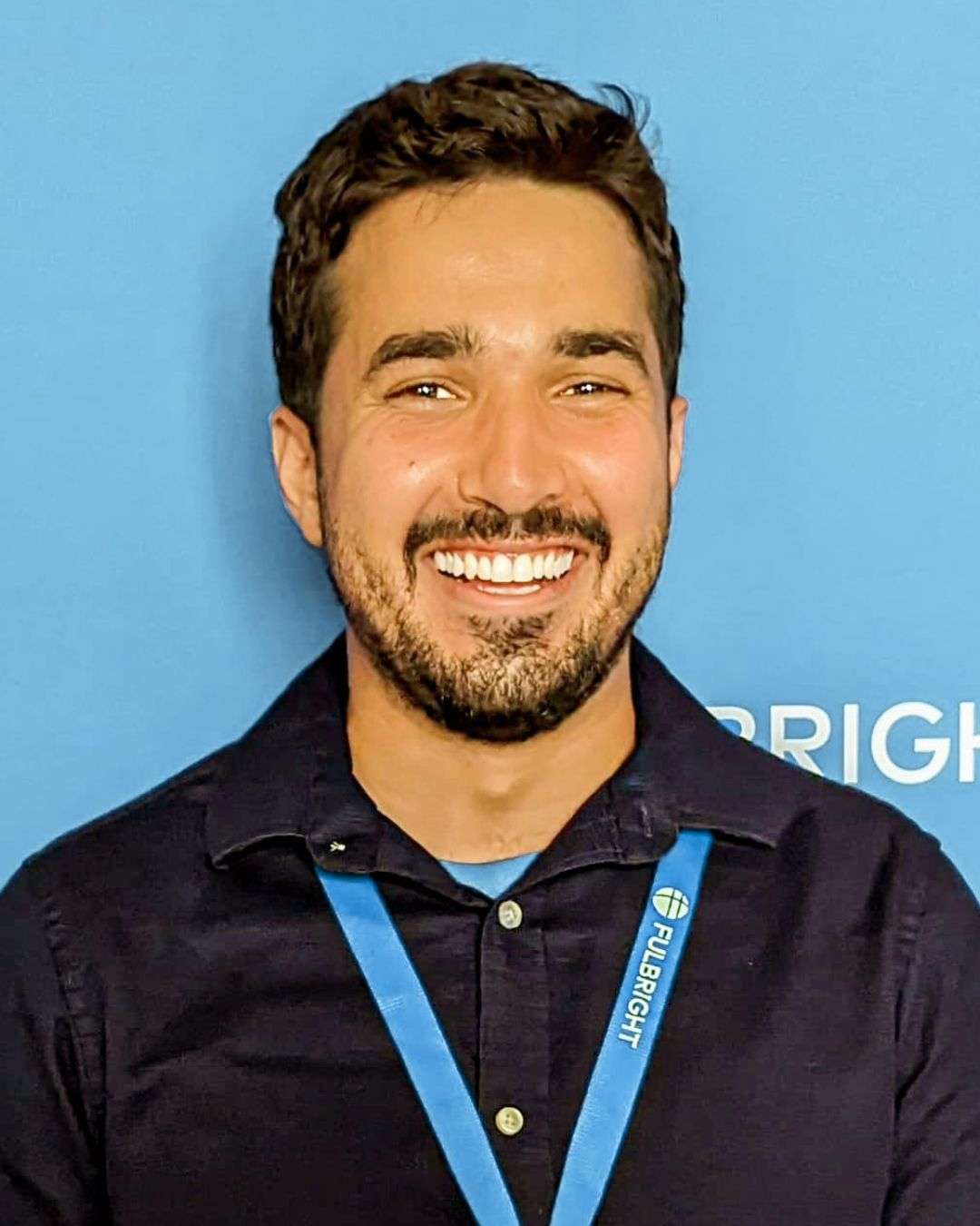 homem sorridente com barba curta usando camisa escura e crachá corporativo, retrato profissional em fundo azul institucional.