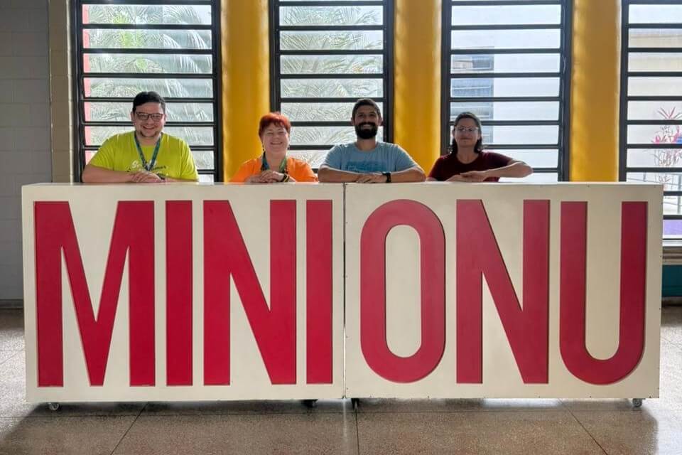 inserir aqui Quatro representantes do Colégio Santa Maria Minas e da PUC Minas posam atrás de um grande painel com a inscrição “MINI ONU”, durante reunião de planejamento do MiniONU Santa Maria Minas 2026, em um espaço interno iluminado com janelas ao fundo.