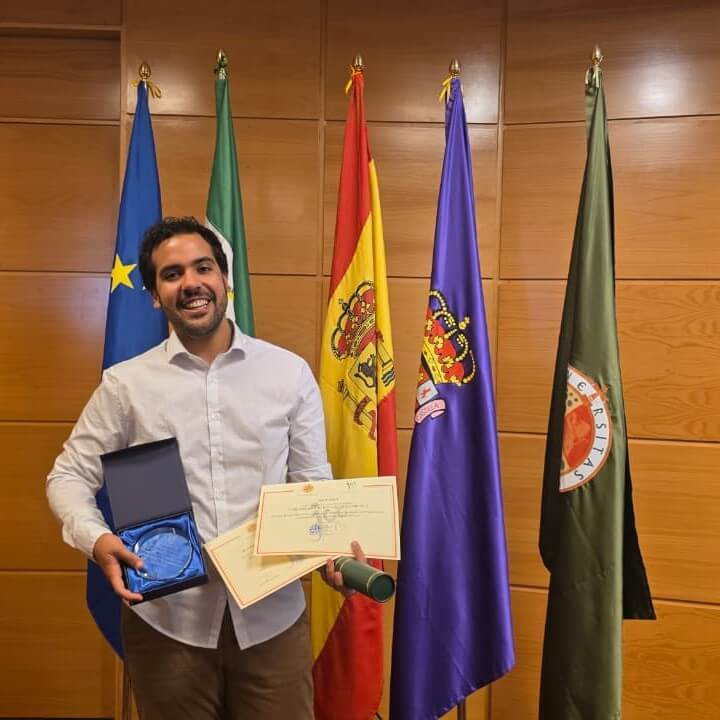 inserir aqui Ex-aluno do Colégio Santa Maria Minas posa sorrindo com medalha, certificados e diploma em cerimônia na Universidade de Jaén, diante de bandeiras institucionais.
